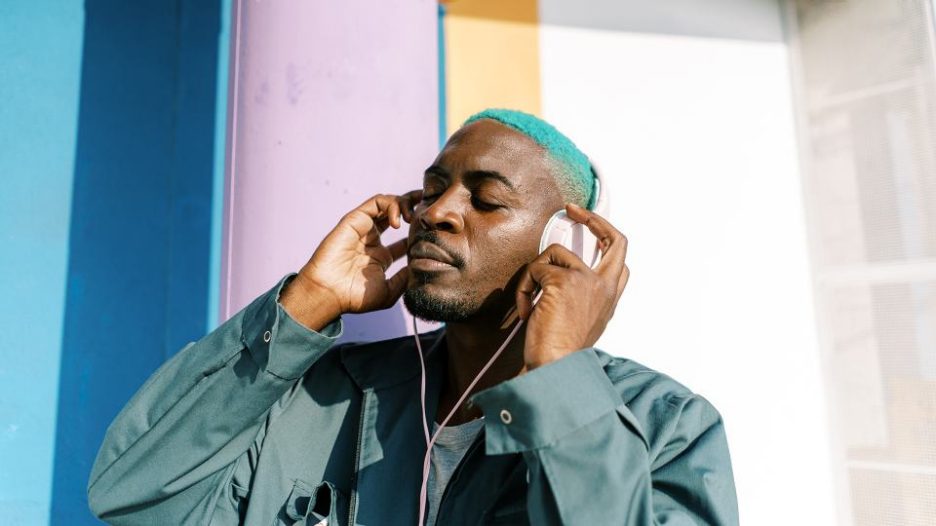 Relaxed African American male with closed eyes wearing trendy outfit standing on street and listening to music in headphones