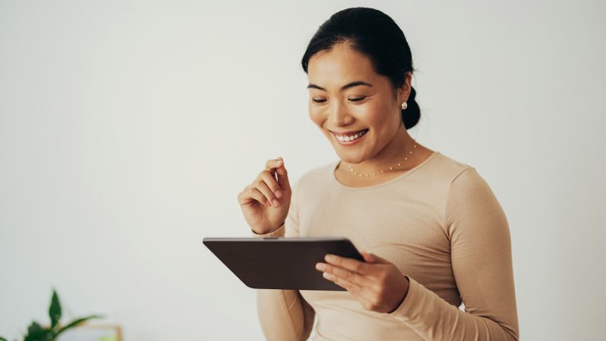 woman in a beige top against a cream wall is smiling at her tablet