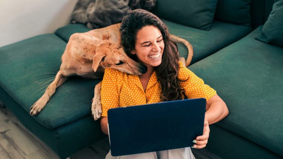 young woman working from home on her laptop smiling as her dog nuzzles into her neck