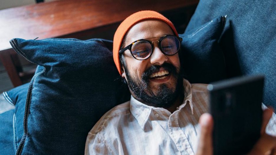 A from above view of a smiling Indian influencer fashionably dressed spending his leisure time relaxing using his smartphone.