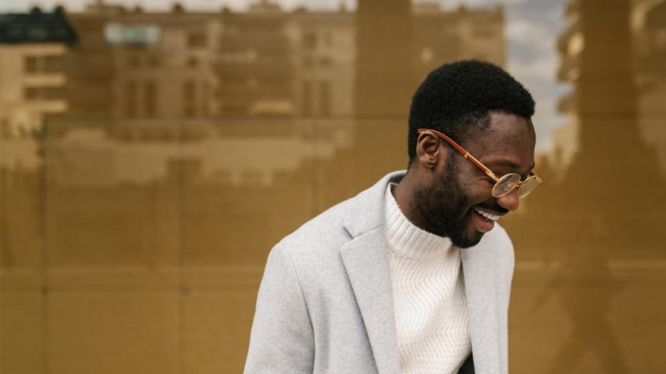 cheerful young bearded male manager in smart casual outfit and eyeglasses smiling brightly on street against building with glass walls