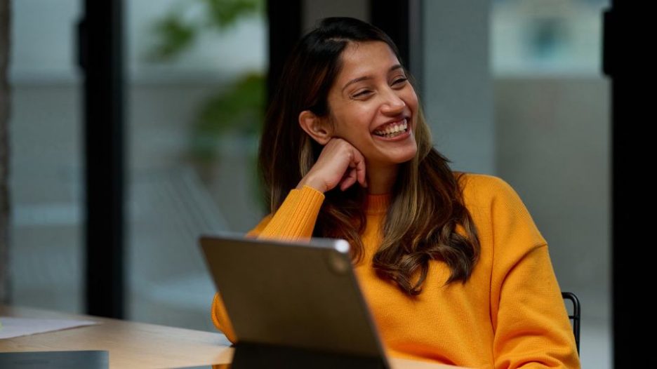 female worker in bright pullover laughing at her desk in office and chatting with anonymous co-worker.