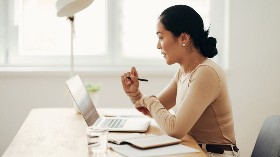 woman in a beige top is looking at her laptop while seated in a light filled office