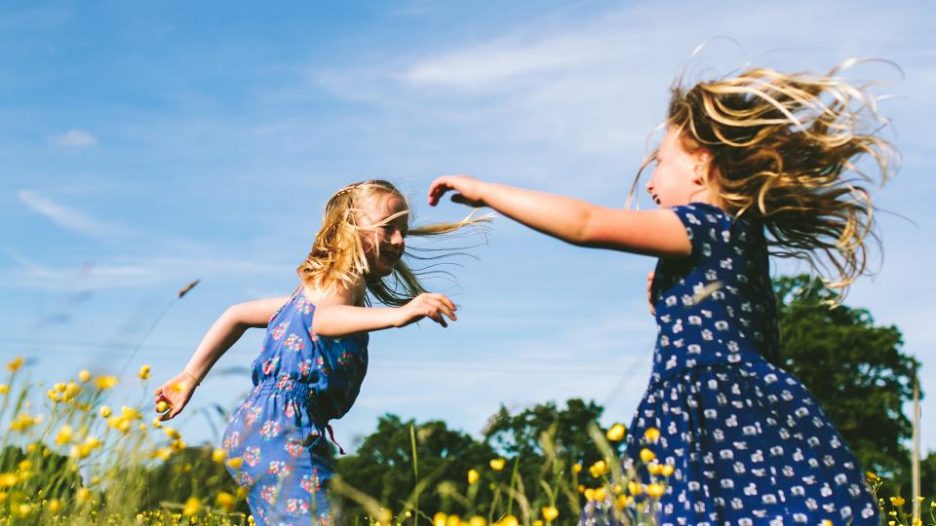 2 little girls dancing in a wildflower meadow having the best fun. Their long blonde hair is blowing in the breeze and they are smiling and shrieking with laughter. Concept of childhood joy.