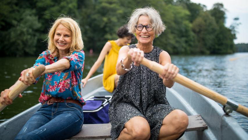 two mature women rowing a boat on the water with a third woman in the background