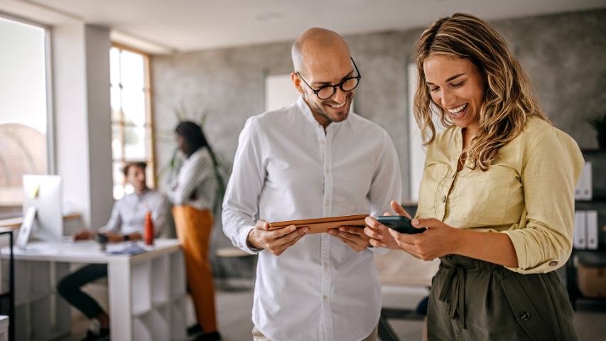 male and female coworker standing in a modern ofice and smiling while using a tablet and a phone