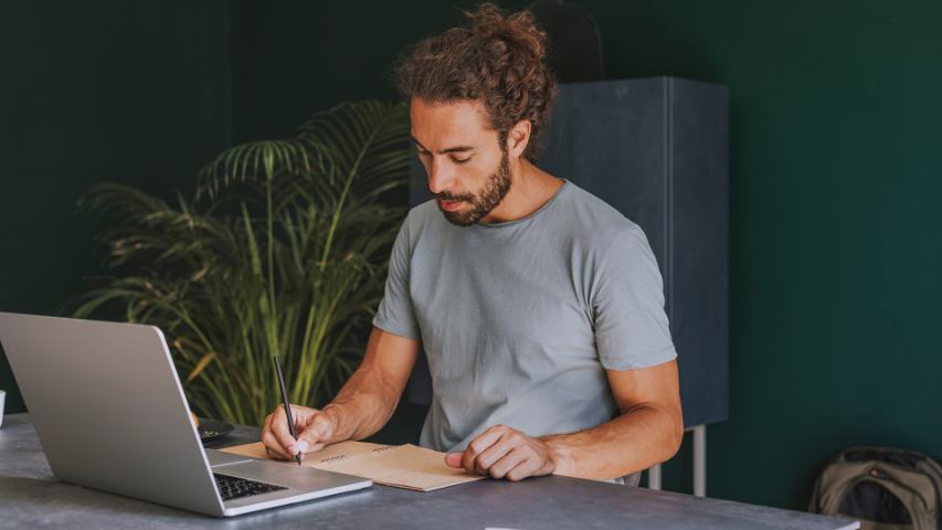man in a grey tee shirt on a laptop in an office with a green wall behind