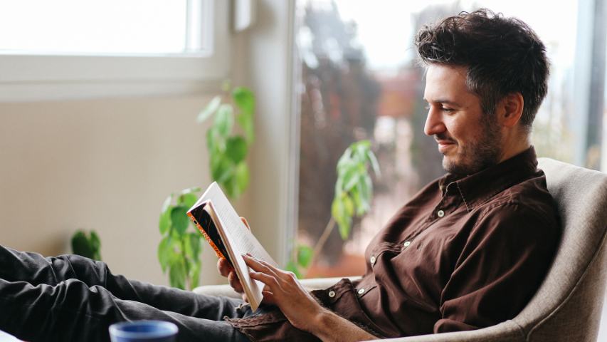 man in a brown shirt is relaxing with his feet up with a book and a hot drink