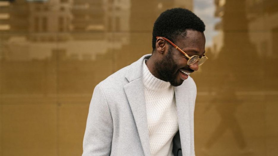 cheerful young bearded male manager in smart casual outfit and eyeglasses smiling brightly on street against building with glass walls