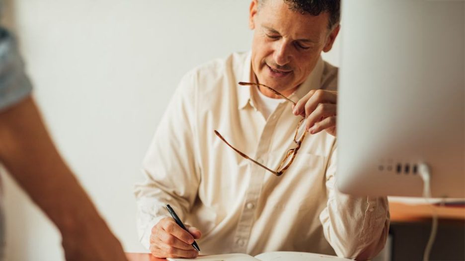 man holding glasses and writing with pen at desk in office