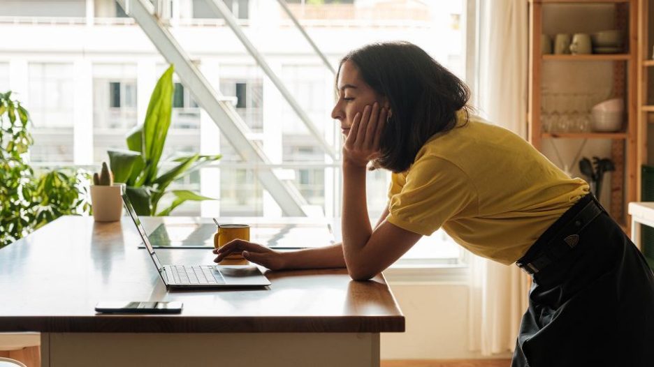 Side view of a young woman working on laptop in kitchen at home