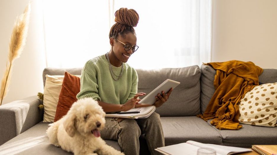 Focused cute stylish African American female student, studying remotely from home, digital tablet, taking notes on notepad during online lesson, e-learning concept. She has a beautiful pet dog by her side