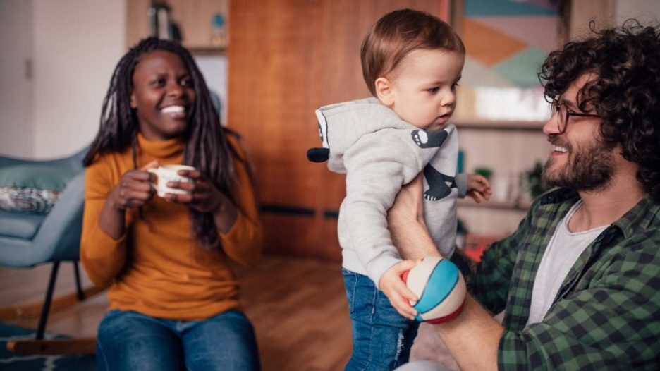 a happy man holding an uncertain toddler while a smiling woman looks on