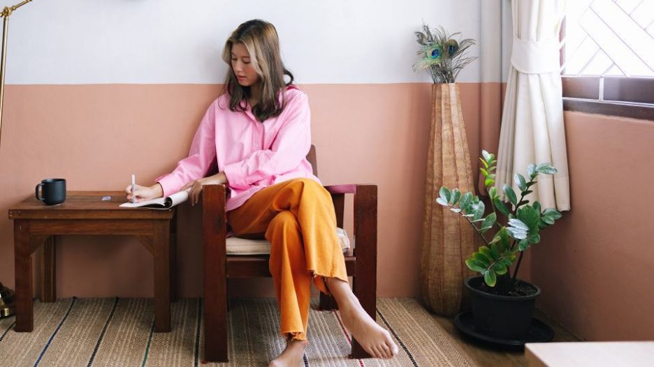 woman in a pink shirt writing in a notebook while sitting on a wooden chair at home