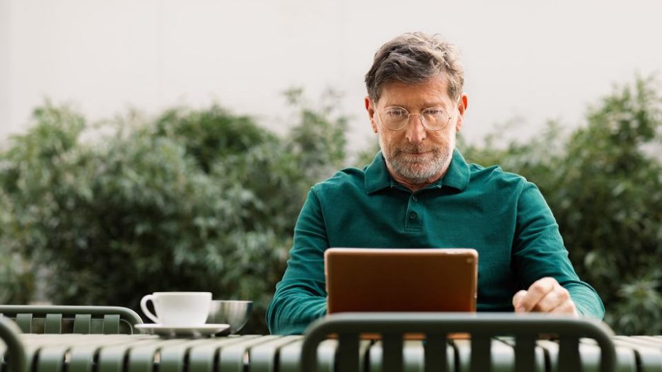 senior man sitting outdoors on terrace, drinking coffee and using digital tablet