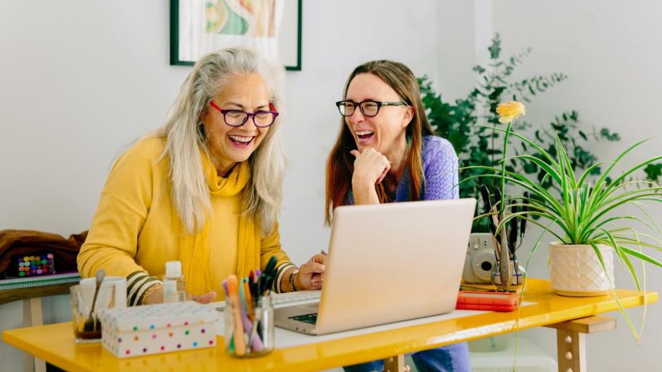 two mature female colleagues laughing together as they work together at one desk