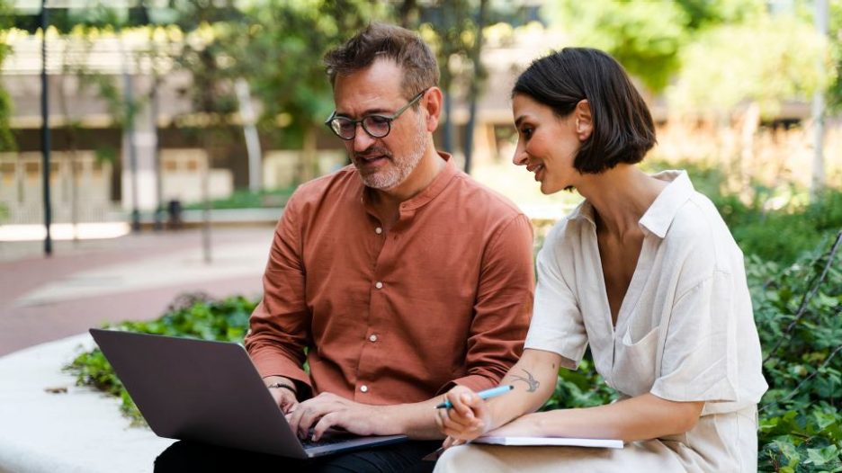 businessman and businesswoman sitting on a park bench working on laptop