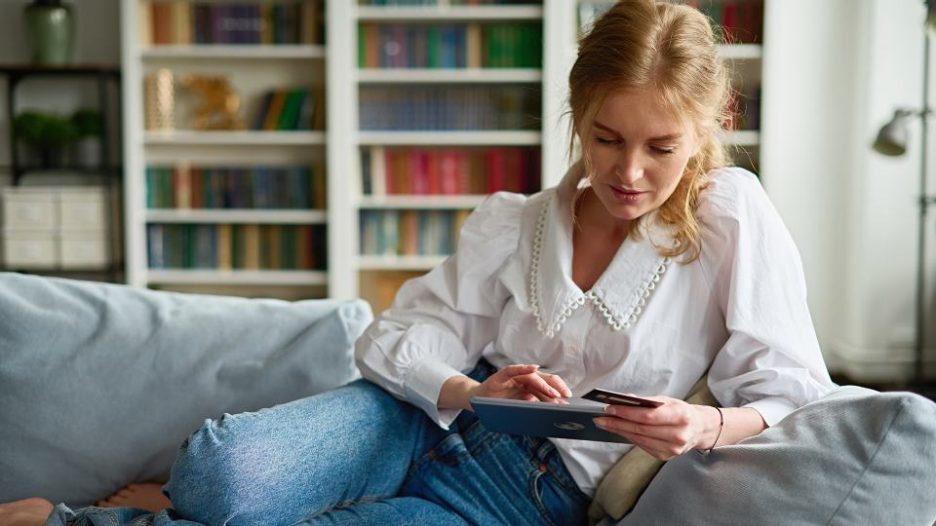 focused female with plastic card making online payment on tablet while sitting on couch in light living room at home