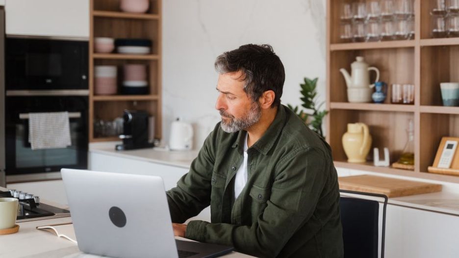 middle-aged bearded male  sitting in the kitchen reviewing bank accounts using laptop and taking notes in notebook